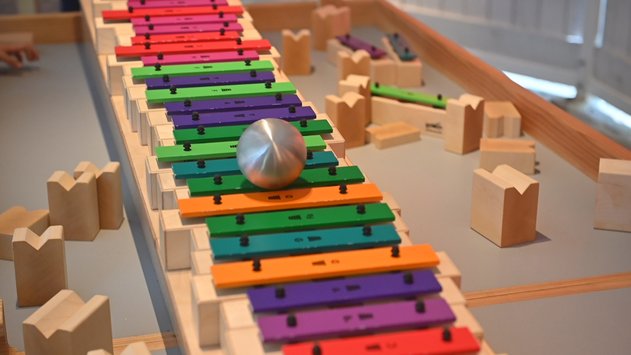 Ein Klangxylophon, das aus bunten, aneinandergereihten Metallplättchen besteht. Auf diesen Klangplatten rollte gerade eine Metallkugel entlang.
