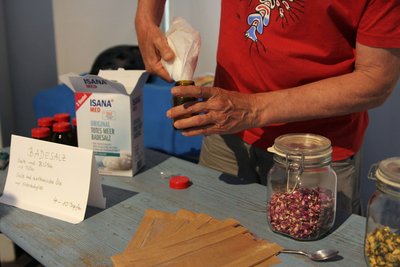 Eine Person steht an einem Tisch und füllt mit beiden Händen etwas aus einem braunen Papiertütchen in eine kleine braune Flasche. Auf dem Tisch stehen Glasgefäße mit getrockneten Blüten und Kräutern.