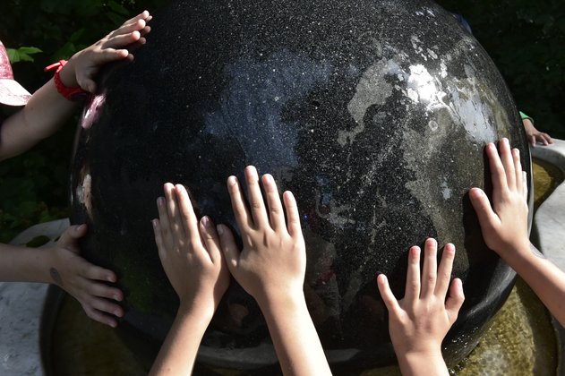 Mehrere Kinderhände berühren eine große, dunkle Kugel aus Granit, die in einem Steinbecken liegt. Im Becken ist ein Film aus Wasser, der dafür sorgt, dass die Kugel gedreht werden kann.